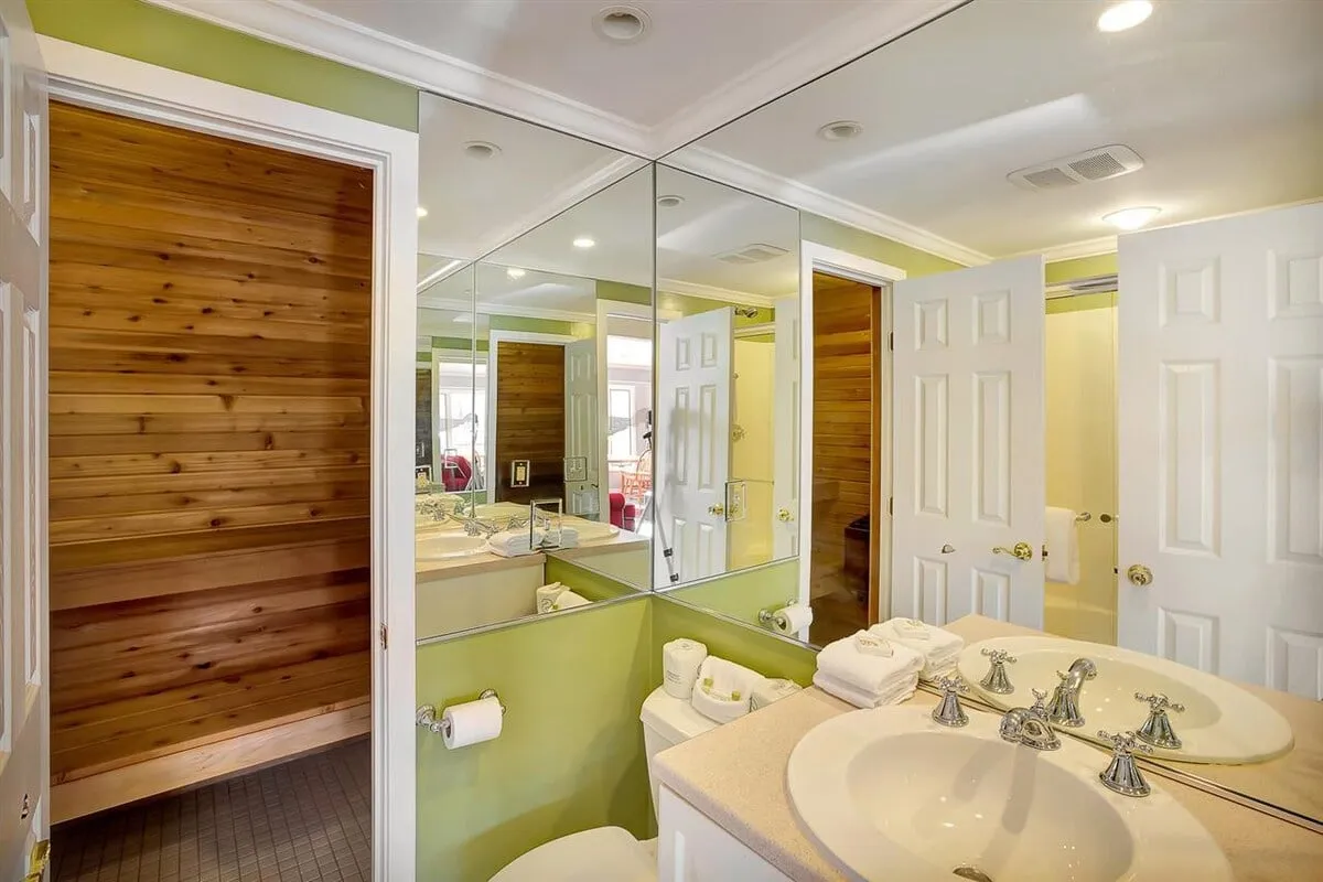 This interior shot features dual sinks, mirrored walls, and a wooden sauna door, suggesting a spa-like atmosphere. The sauna is subtly visible, inviting guests to imagine the soothing experience it offers after a day outside.