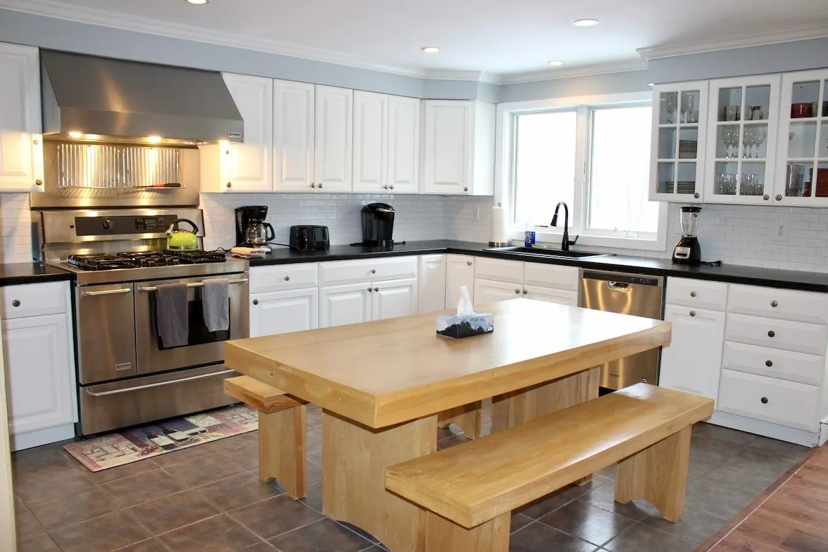 The image showcases a warm, inviting dining area with a large wooden table perfect for group gatherings, complemented by modern stainless steel appliances and bright white cabinetry. Sunlight streams in through a window, illuminating the communal space and creating a cozy, welcoming atmosphere ideal for shared meals and après-ski relaxation.