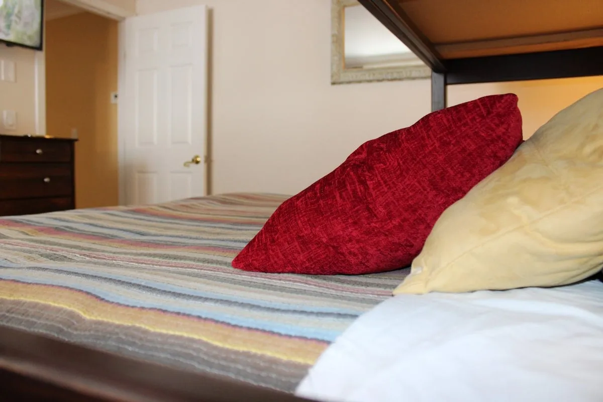 An inviting bedroom view featuring a red decorative pillow, striped bedding, a yellow pillow, and a decorative mirror. The arrangement and color accents create a calm, welcoming atmosphere perfect for restful mornings or intimate evening chats.