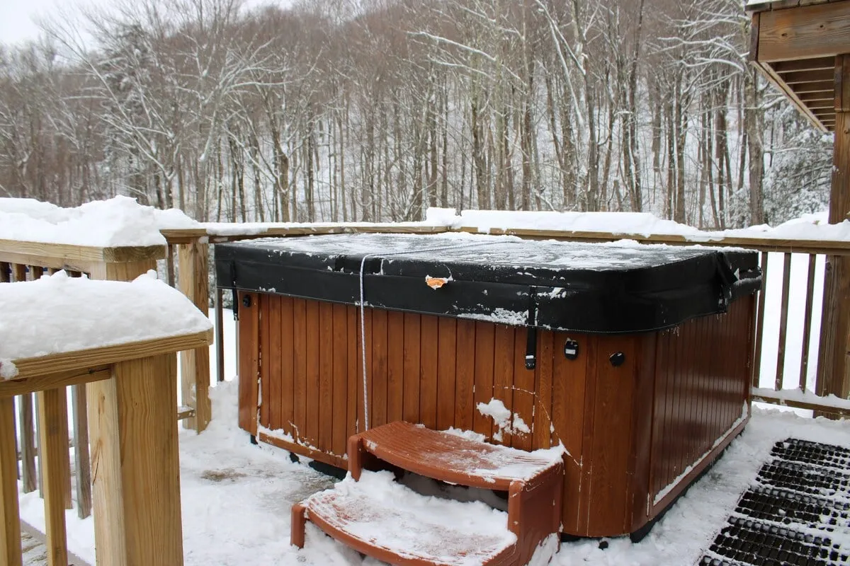 This image displays a hot tub situated on a snow-covered deck, surrounded by trees, creating a welcoming and peaceful environment. The setting evokes the relaxation of soaking in the hot tub after a day of outdoor activities, with the warmth of the tub contrasting against the crisp mountain air and snowy landscape.