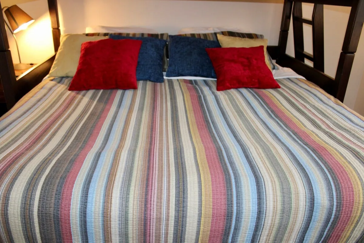 A cozy king bedroom with a striped bedspread, colorful pillows, and a soft glow from the bedside lamp. The setting suggests a comfortable space ideal for relaxing, reading, or settling in for a restful night.