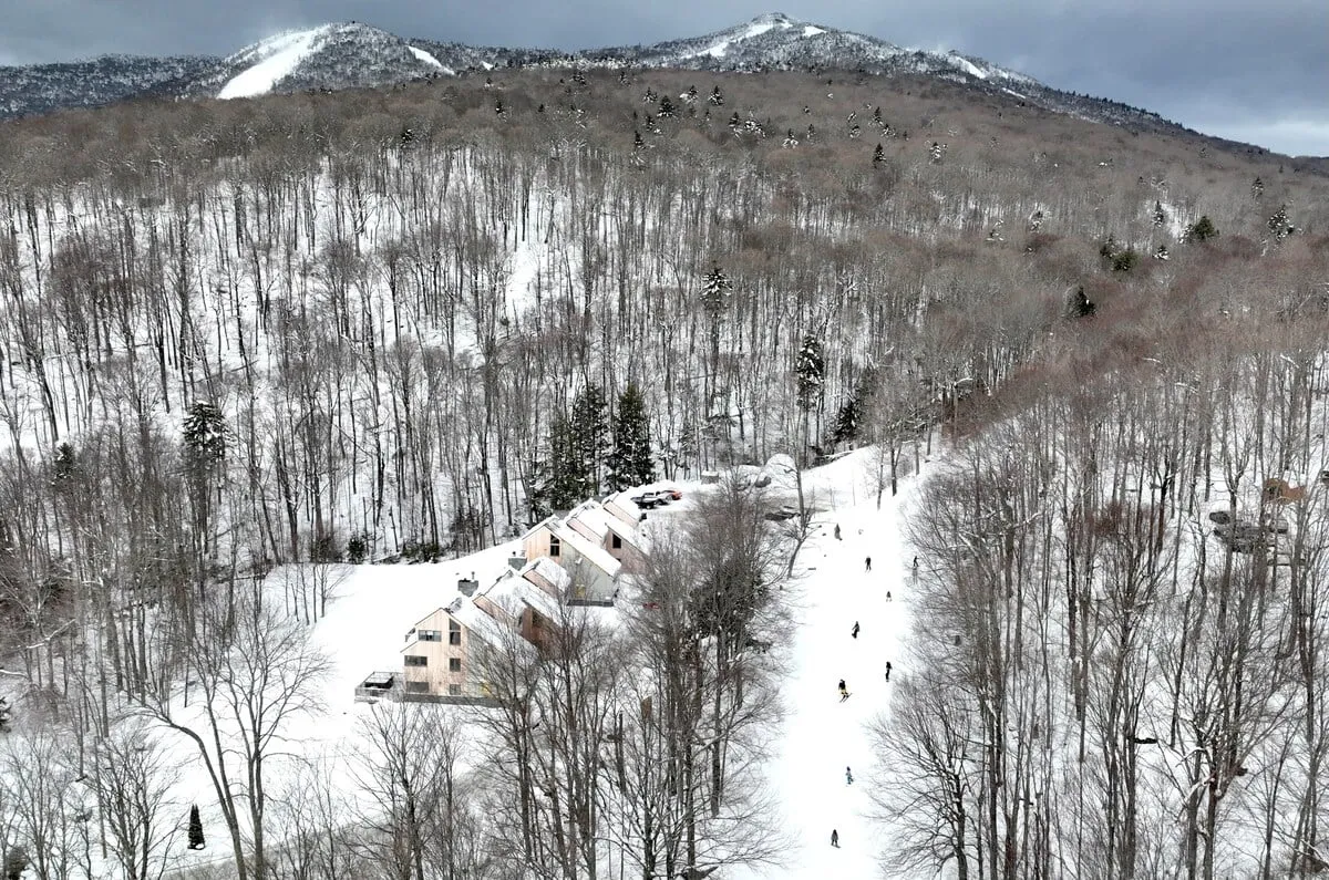 A snow-covered landscape featuring the property structure set against picturesque mountains. The immediate area shows pristine snow, ski tracks, and a wintery atmosphere, clearly indicating direct access to the slopes. The serene ambiance highlights the excitement and convenience of stepping right out to the trails.